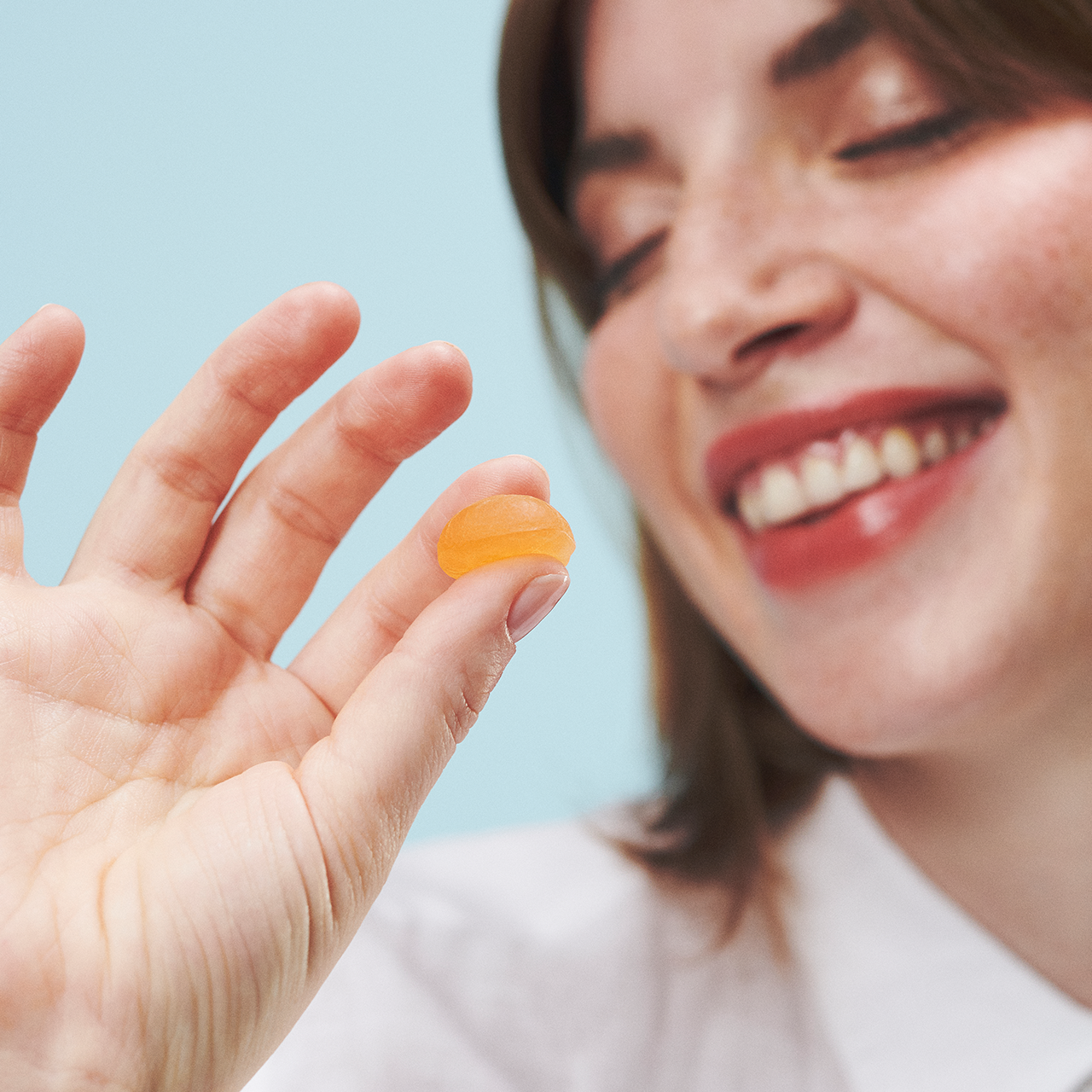 A closeup shot of a female model smiling, eyes closed, with her hand holding a Feals Relax Gummy set against a light blue background.